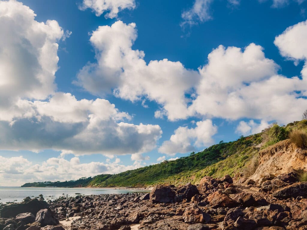 Staning on the beach looking over the rocks towards Sunnyside North Beach. Green cliffs in the background and blue skies with puffy white clouds