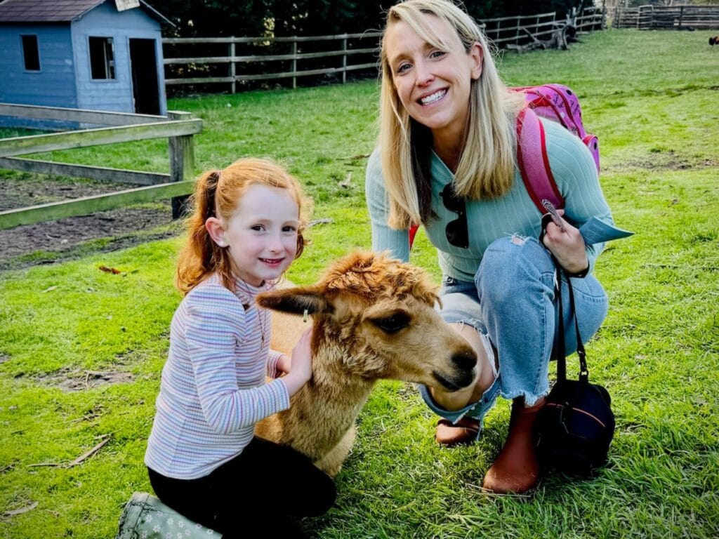 A mother and daughter in a paddock squatting down to pat an Alpacce.