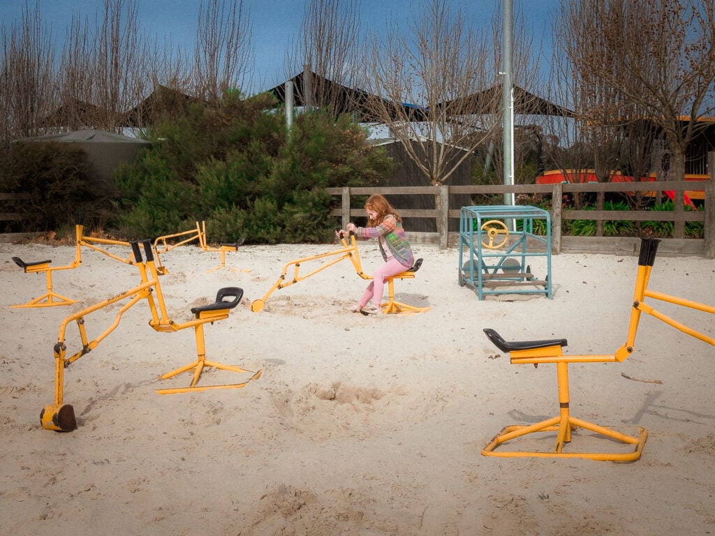 A five year old in the middle of a huge sand pit using a toy sandpit digger. 