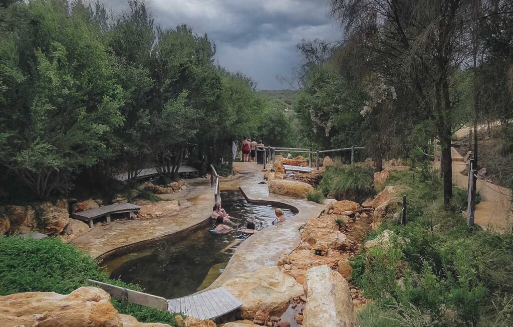 View of a hots springs pool made of earthy sandstone, surrounded by trees with a view into the bush on an overcast winter's day.