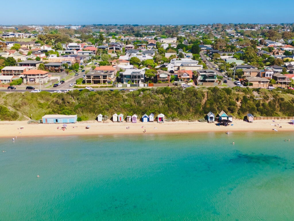 Aerial view looking straight onto Fishermans Beach. Crystak aquamarine water in the foreground and a long stretch of beach dotted with colourful bathing boxes.