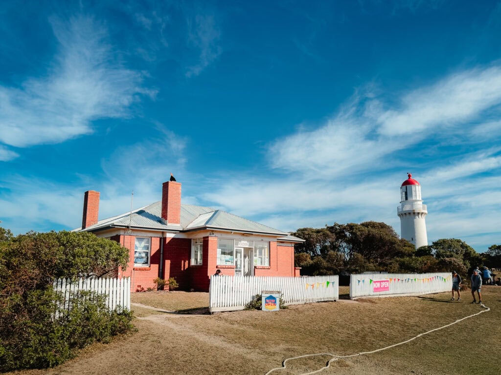 Old red brick Kiosk building with a white picket fence. The lighthouse is standing in the background surrounded by trees.
