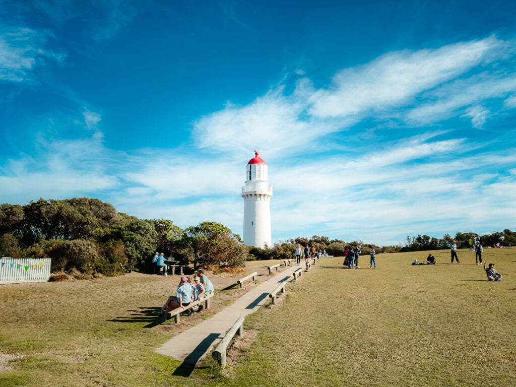 Cape Schanck Lighthouse with its white walls and bright red roof standing tall at the end of a path, surrounded by green lawn with people walking on it.