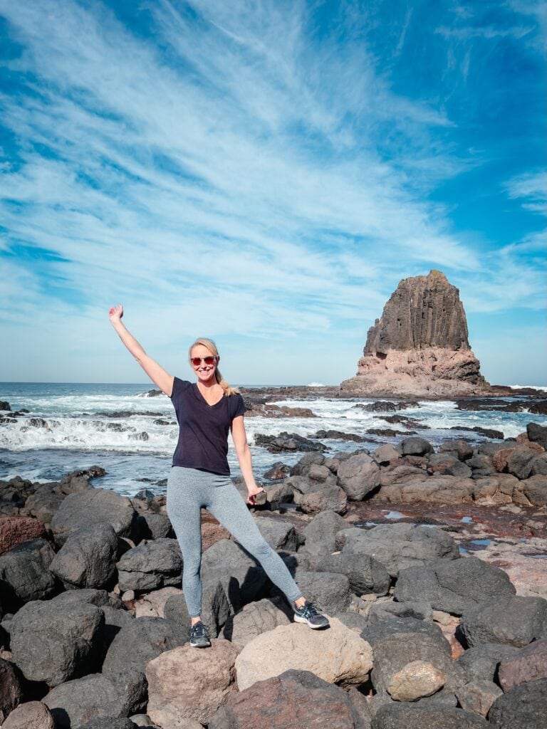 Me standing on a black stone beach with the large volcanic Pulpit Rock in the background. The ocean is swirling around the base of the rock.